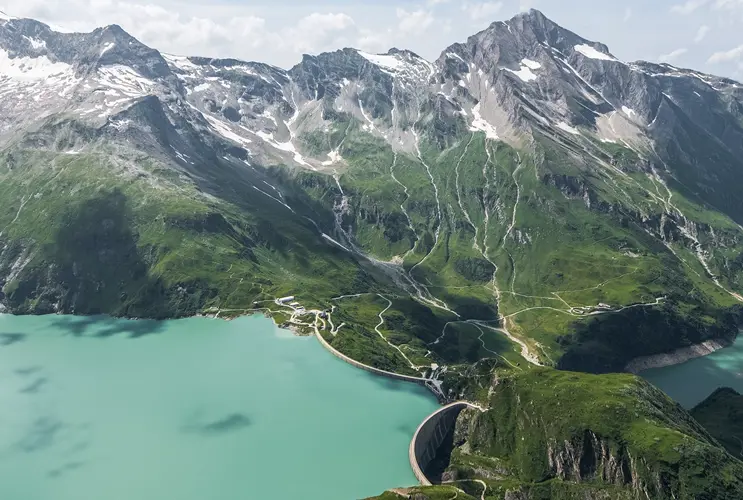 Wir sehen zwei Stauseen in hellem türkis getrennt durch eine Staumauer. Dahinter Hochgebirge im Sommer. Der hellblaue Himmel wird von der Sonne erstrahlt.